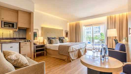 Hotellrum med dubbelsäng, sittgrupp och kök, samt balkong med utsikt. (AI-genererad text)