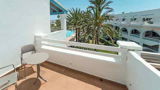 En balkong med bord och stolar, med utsikt över palmer och en pool. (AI-genererad text)