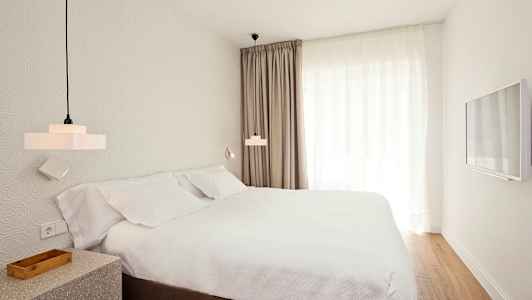 Rum med en säng, vita lakan och gardiner, samt väggmonterad TV. (AI-genererad text)