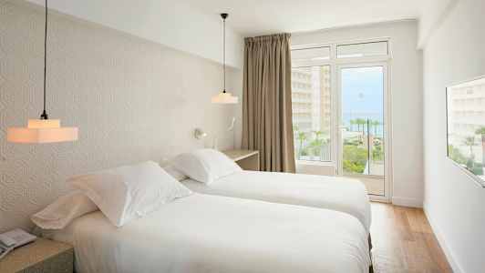 Ett hotellrum med två enkelsängar, ljus inredning och en balkong med utsikt mot havet. (AI-genererad text)