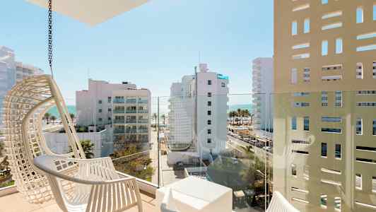 En balkong med vita stolar och utsikt över moderna byggnader samt havet i bakgrunden. (AI-genererad text)