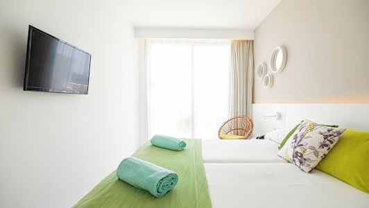 Ett ljust hotellrum med en dubbelsäng, färgglada kuddar, hoprullade handdukar och en platt-TV på väggen. (AI-genererad text)