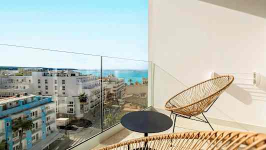 En balkong med två stolar i rotting och ett runt bord, med utsikt mot havet och stadens byggnader. (AI-genererad text)