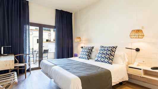 Ett hotellrum med en dubbelsäng, blåa dekorkuddar och nattlampor. Ett skrivbord med stol står vid sidan om, samt en balkong med bord och stolar. (AI-genererad text)