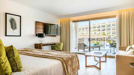 Ett hotellrum med en säng, soffgrupp och balkong med utsikt över poolområdet och hotellet. (AI-genererad text)