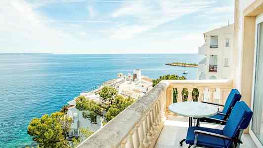 Utsikt från en balkong med bord och stolar över havet och omgivande byggnader. (AI-genererad text)