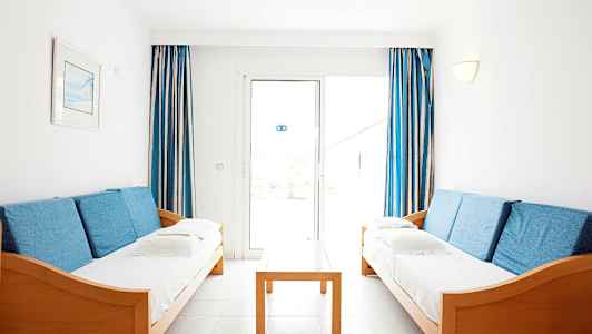 Två soffor med blå kuddar står mittemot varandra, med ett träbord emellan och blåvita gardiner vid fönstret i bakgrunden. (AI-genererad text)