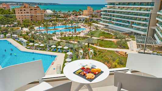 Utsikt från en balkong mot en hotellpool och havet, med en fruktbricka och två glas på ett bord. (AI-genererad text)