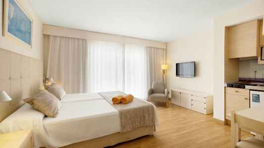 Ett hotellrum med dubbelsäng, två handdukar på sängen, en fåtölj, en väggmonterad TV och en liten kökshörna. (AI-genererad text)