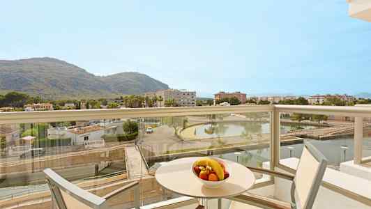 En balkong med utsikt över ett landskap som inkluderar berg, byggnader och en damm. På bordet finns en skål med frukt. (AI-genererad text)