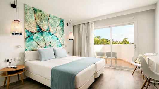 Ett hotellrum med en dubbelsäng, blå sängkläder, en färgglad tavla ovanför sängen, och en balkong med två stolar utanför. (AI-genererad text)