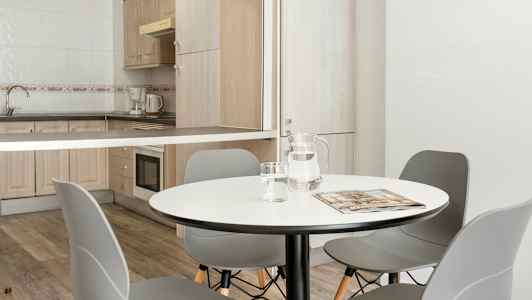 En matsal med ett runt bord, grå stolar och en angränsande köksyta med träskåp och vitvaror. (AI-genererad text)