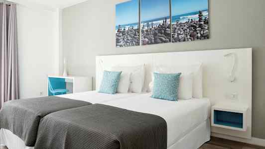 Ett hotellrum med två enkelsängar, vita sängkläder och blå prydnadskuddar. På väggen hänger en tredelad tavla föreställande en stenstrand. (AI-genererad text)