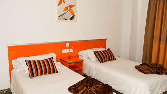 Två enkelsängar med vita lakan, bruna filtar och randiga kuddar i ett rum med orange inredning. (AI-genererad text)