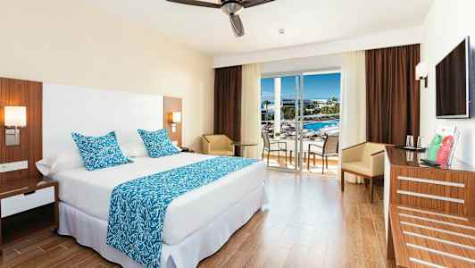 Ett hotellrum med en stor säng, blå dekorativa kuddar och täcke, samt ett bord med stolar vid ett glasfönster som leder till en balkong. (AI-genererad text)