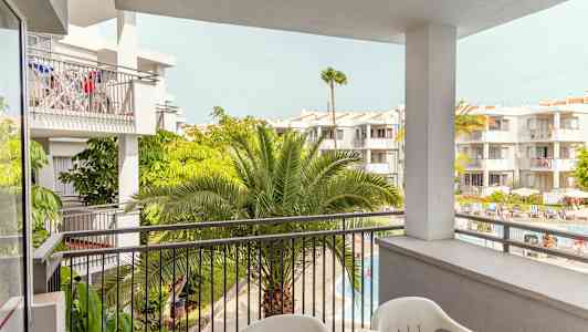 Utsikt från en balkong mot flera vitmålade byggnader med grönskande palmer och en poolområde nedanför. (AI-genererad text)