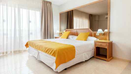 Ett hotellrum med en dubbelsäng, vita och gula sängkläder. En stor spegel bakom sängen och ljusa gardiner vid fönstret. (AI-genererad text)