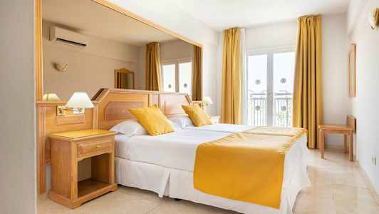 Ett hotellrum med en dubbelsäng, vita lakan och gula kuddar och överkast. Rummet har ljust trämöblemang och fönster med gardiner. (AI-genererad text)