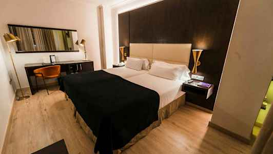 Ett hotellrum med en dubbelsäng, mörka och ljusa färgtoner på möbler samt en skrivbordshörna med stol och spegel. (AI-genererad text)