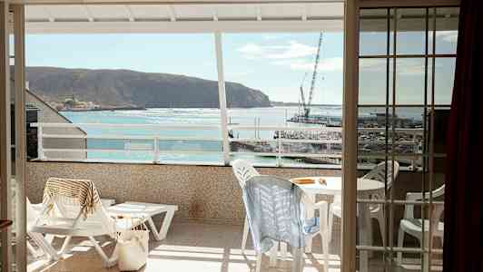 Utsikt från en balkong mot havet och en liten hamn, med utemöbler och solstolar på balkongen. (AI-genererad text)