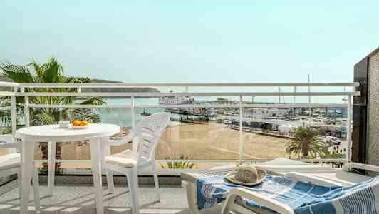 Vy från en balkong med vita utemöbler, med utsikt över en strand, en småbåtshamn och havet i bakgrunden. (AI-genererad text)