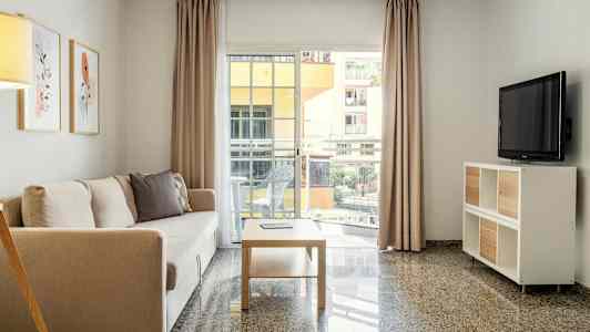 Ett vardagsrum med en soffa, ett soffbord och en TV samt stora fönster som vetter mot en balkong. (AI-genererad text)