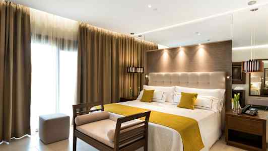 Hotellrum med stor säng, ljusa väggar och gardiner samt en sittbänk vid fotändan. (AI-genererad text)