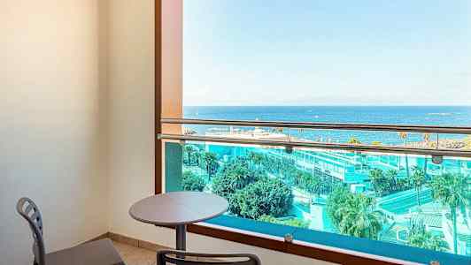 En balkong med bord och stolar, utsikt över havet och hotellområde. (AI-genererad text)