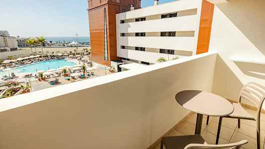 Utsikt från en balkong mot en poolområde med solstolar, parasoller och palmer, samt vita och röda byggnader och havet i bakgrunden. Två stolar och ett bord syns på balkongen. (AI-genererad text)
