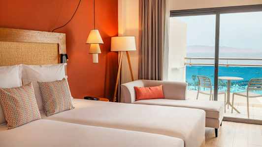 Ett hotellrum med dubbelsäng, dekorativa kuddar, en soffa och glasdörrar som leder till balkongen med utsikt över havet. (AI-genererad text)