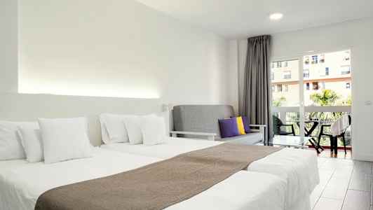 Ett hotellrum med två enkelsängar med vita sängkläder, en grå soffa med färgade kuddar och en balkong med utsikt. (AI-genererad text)