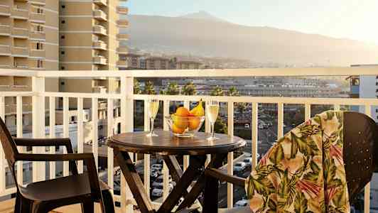 En balkong med ett bord, två glas, en skål med frukt och omgiven av en vy av omkringliggande byggnader och berg. (AI-genererad text)
