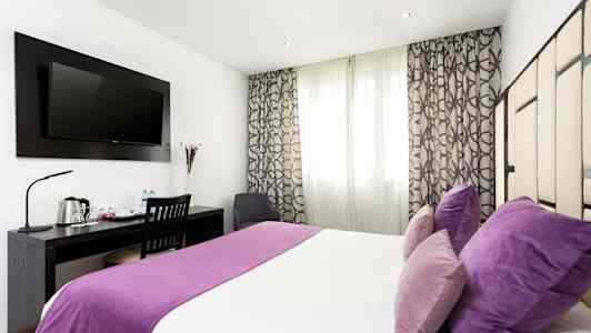 Ett hotellrum med dubbelsäng, lila sängkläder och kuddar, skrivbord med stol och en väggmonterad TV. (AI-genererad text)