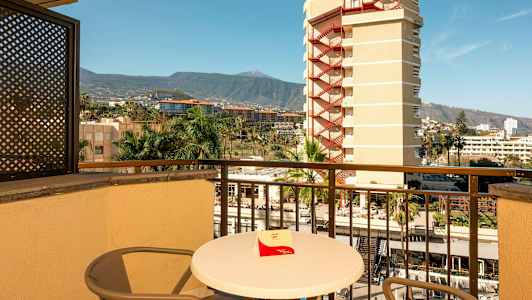 En balkong med bord och stolar, utsikt över omgivande byggnader, palmer och berg i bakgrunden. (AI-genererad text)