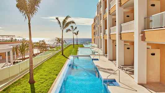 Långsmal pool omgiven av grönska och byggnader med balkonger, med havet i bakgrunden. (AI-genererad text)