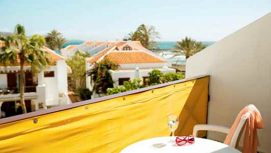 Vy från en balkong med gul avskärmning, ett litet bord med glas och röda solglasögon, vit stol, samt semesterhus och havet i bakgrunden. (AI-genererad text)