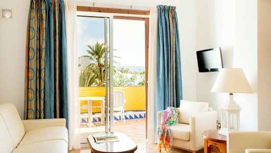 Ett ljust vardagsrum med vita möbler och en öppen balkongdörr mot utsikt med palmer. (AI-genererad text)