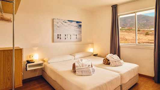 Ett ljust hotellrum med en dubbelsäng, fönster med utsikt över landskapet och en väggmålning ovanför sängen. (AI-genererad text)