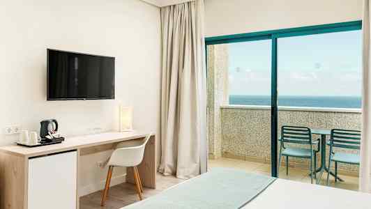Interiör av ett hotellrum med en säng, skrivbord, TV och en balkong med utsikt över havet. (AI-genererad text)