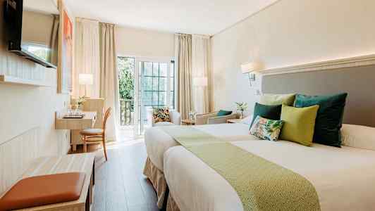 Ett ljust hotellrum med två sängar, prydnadskuddar och en arbetsstation vid balkongen. (AI-genererad text)