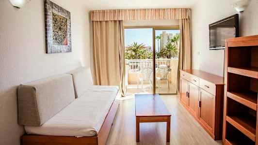 Ett möblerat rum med en soffa, ett bord, en TV och en hylla. Utgång till en balkong med sittmöbler. (AI-genererad text)