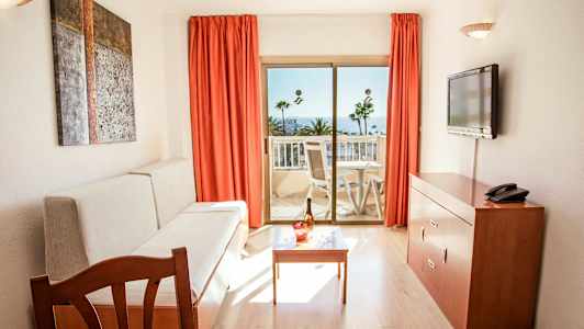 Rum med soffa, bord, TV och balkong med vy mot palmer och hav. (AI-genererad text)