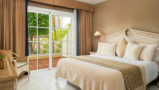 Ett hotellrum med en stor säng, beige sängkläder, vita kuddar och en glasdörr som öppnar mot en terrass med grönska. (AI-genererad text)