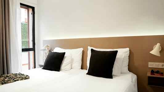 Ett hotellrum med två enkelsängar, vita sängkläder och svarta kuddar, samt ljusa nattlampor på väggen. (AI-genererad text)