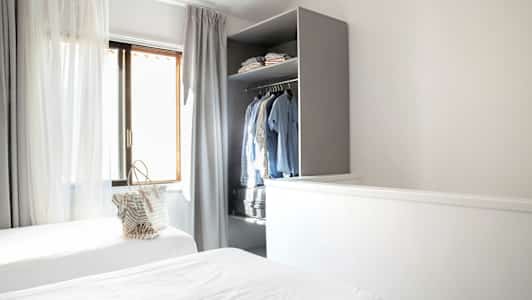 Ett rum med två enkelsängar, gardiner vid fönstret och en öppen garderob med kläder. (AI-genererad text)