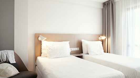 Ett hotellrum med två enkelsängar, ett fönster med gardiner och en stol på sidan. (AI-genererad text)