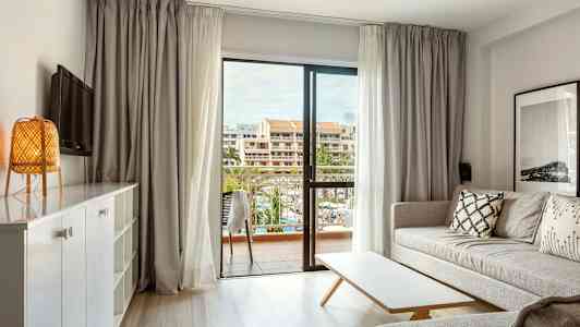 Ljust vardagsrum med soffa, bord och utsikt mot en balkong med byggnader i bakgrunden. (AI-genererad text)