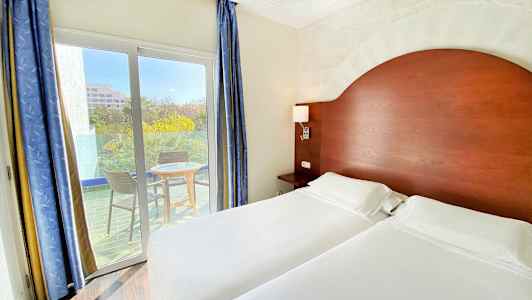 Ett hotellrum med två enkelsängar, ett trähuvudgavel, och en balkong med två stolar och ett bord utanför. (AI-genererad text)