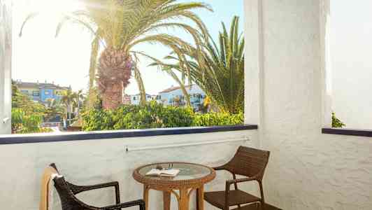 En balkong med två stolar och ett bord med en bok. Bakom balkongen syns en palm och byggnader. (AI-genererad text)