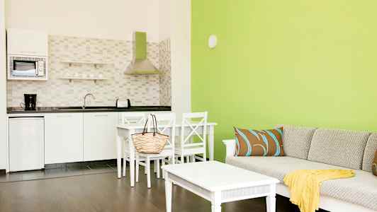 Ett vardagsrum med gröna väggar, en soffa med kuddar och en bord, samt ett angränsande kök med vita skåp och apparater. (AI-genererad text)
